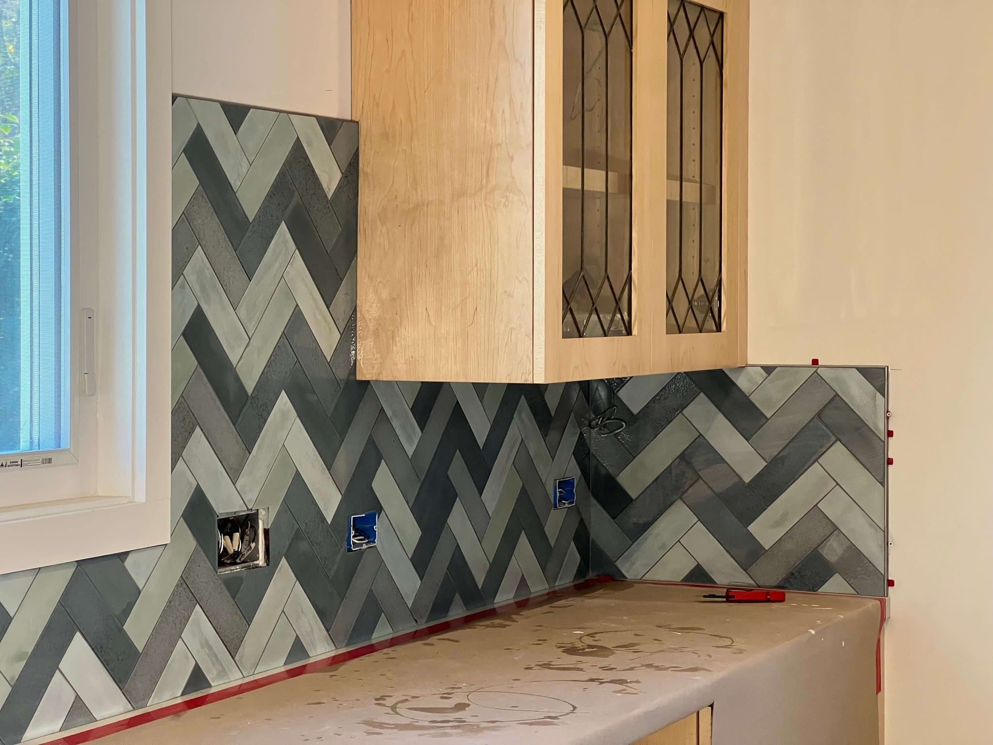 Close-up of a herringbone tile backsplash wrapping a kitchen corner beneath a maple glass-front cabinet